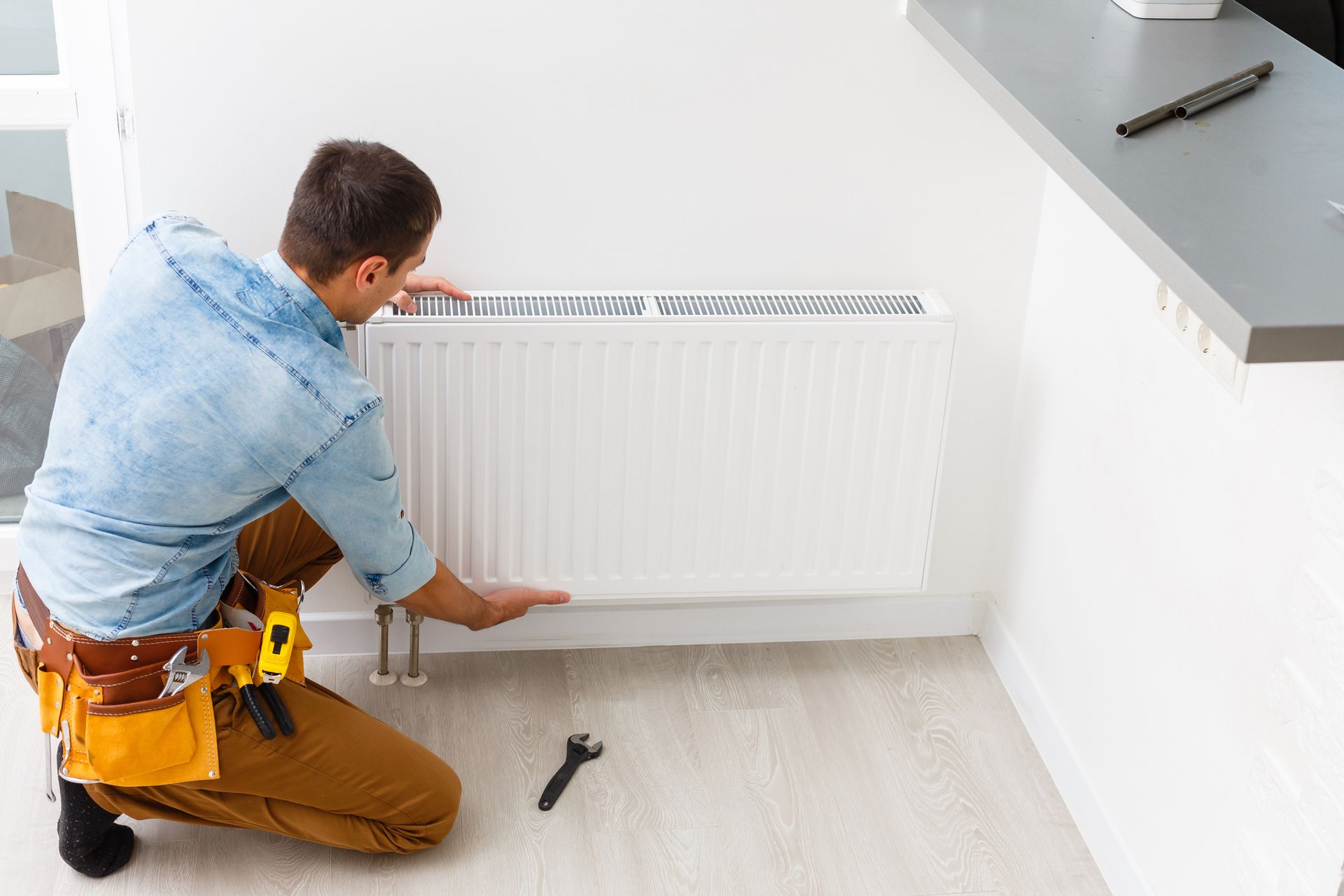A man in work clothes installs a white radiator in a bright room. He kneels, working near a wall and countertop.