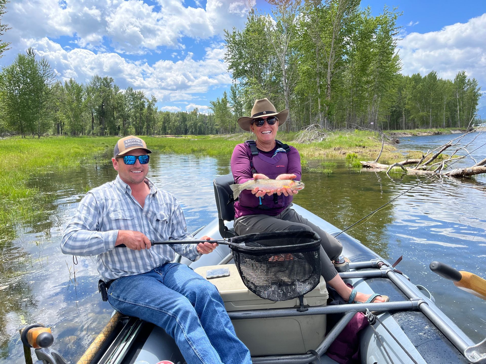 Two people fly fishing from a boat on a lake; one holds a fish, smiling. Green trees and blue sky.