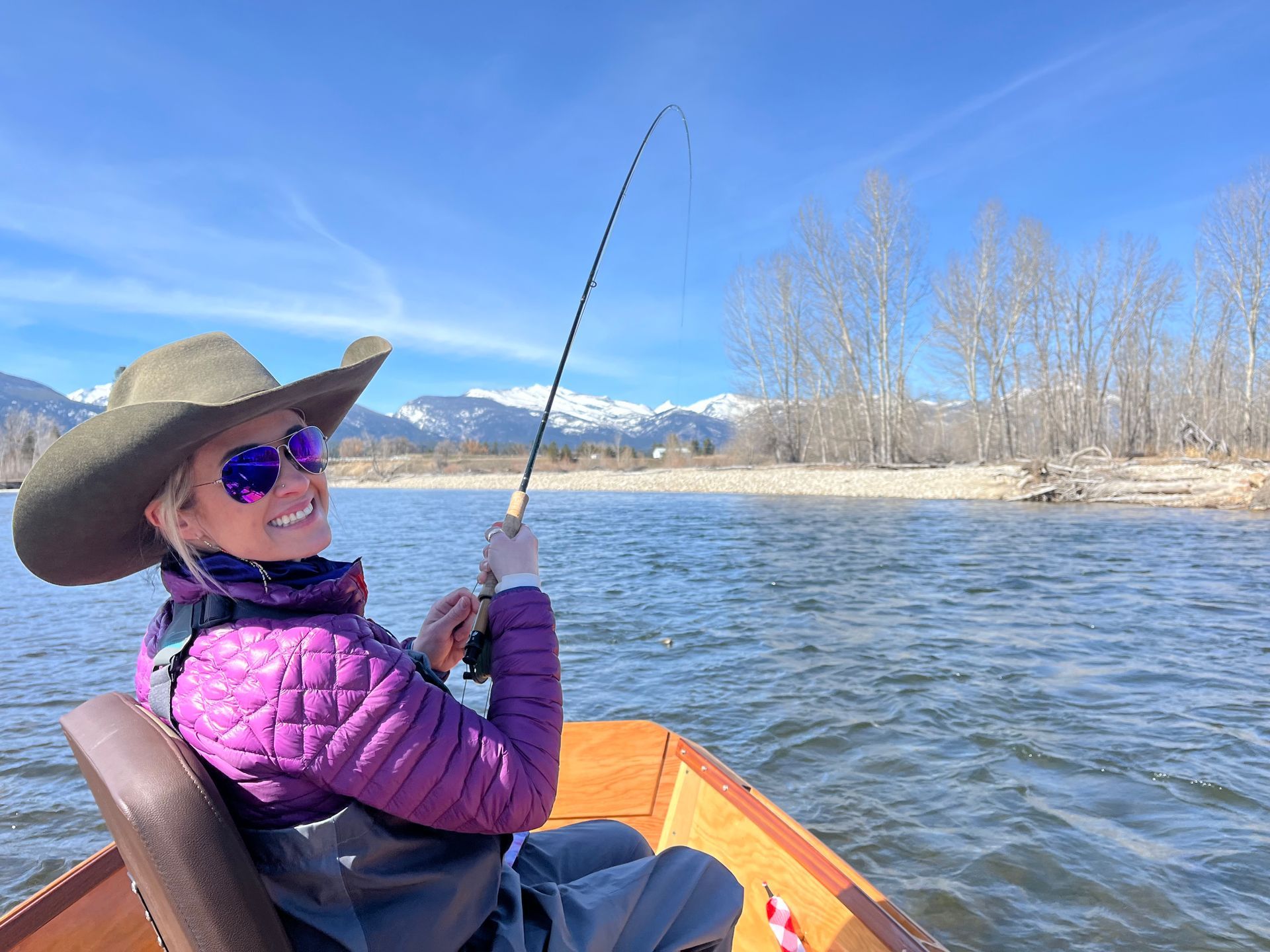Woman fishing from a boat on a river, wearing a hat and purple jacket, mountains in background.