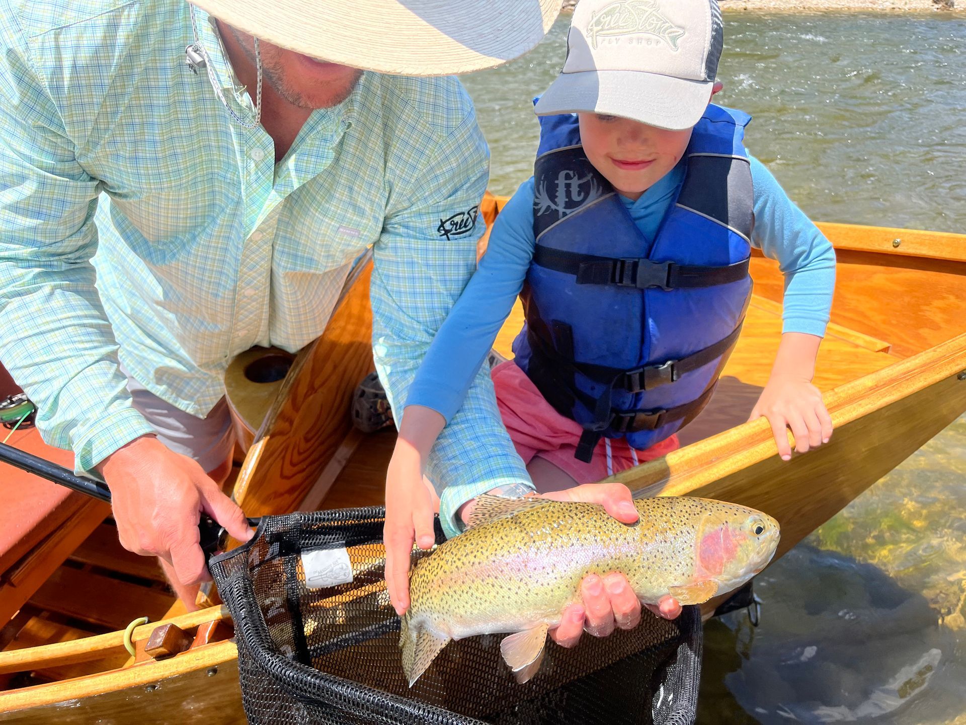 Person helping a child hold a rainbow trout in a net on a wooden boat.