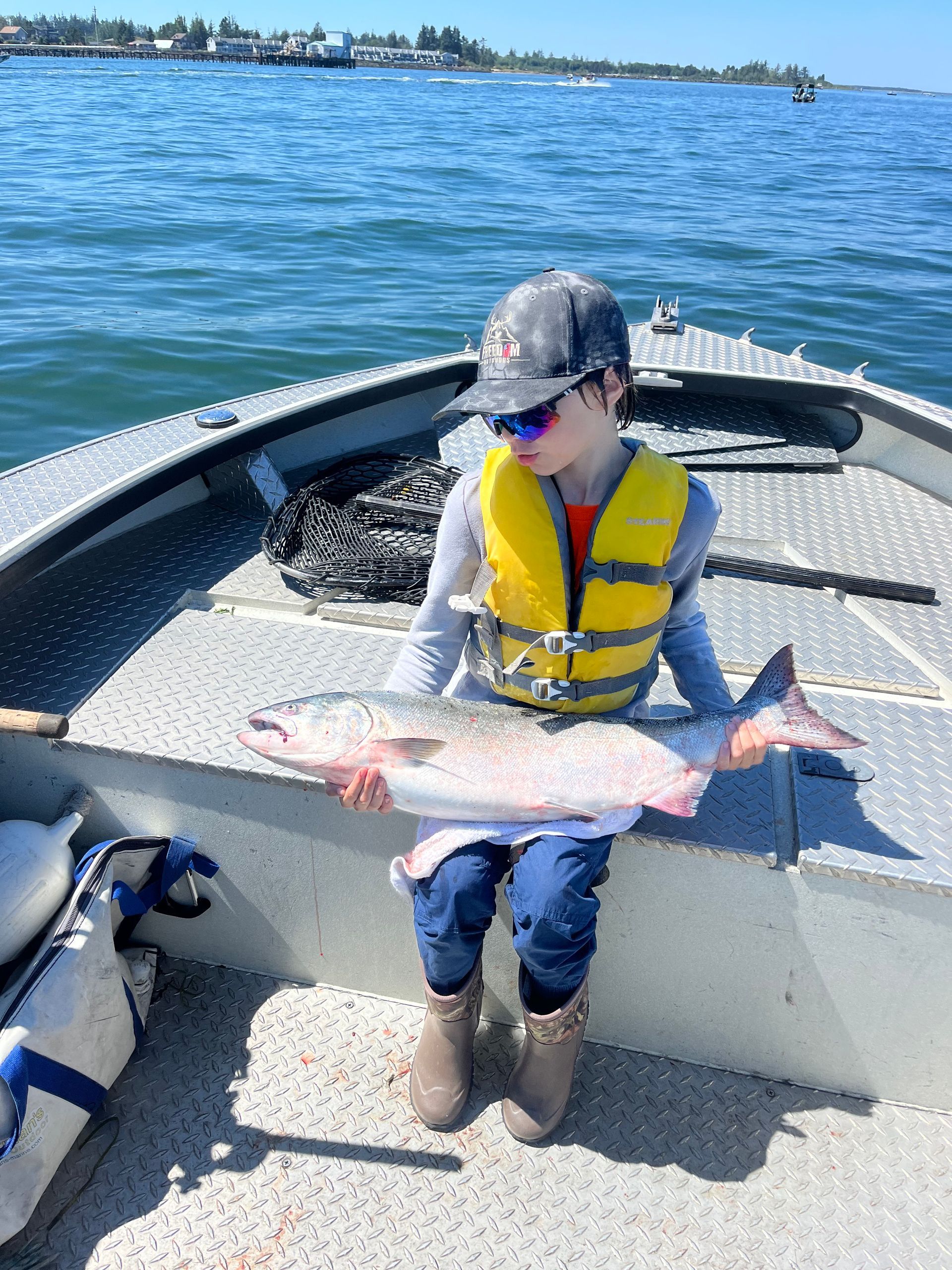 Boy on a boat holds up a large fish, wearing a life vest and sunglasses, on a sunny day.