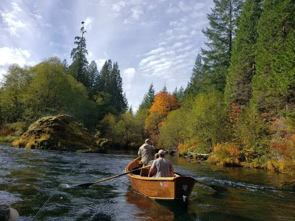 Two people in a wooden drift boat on a river surrounded by trees with fall foliage.