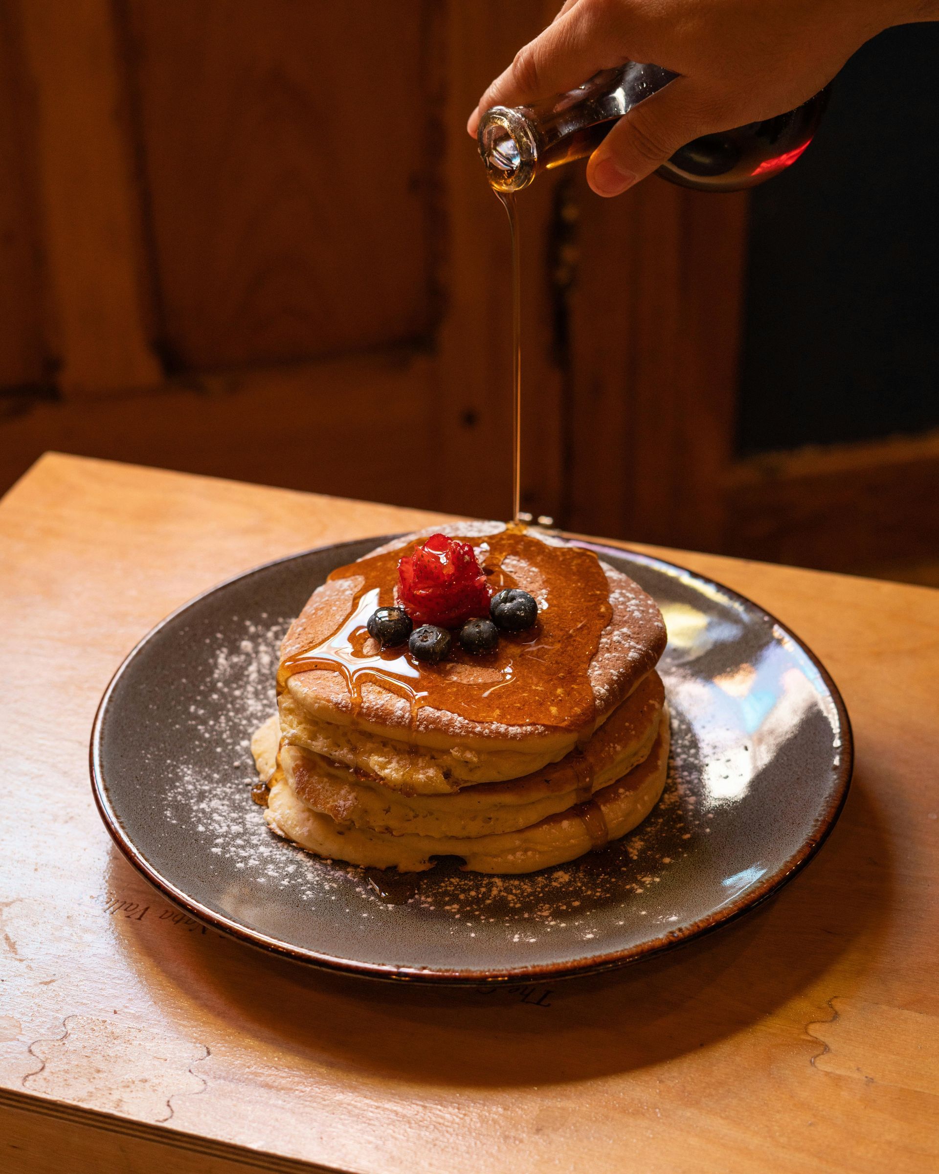 Stack of pancakes topped with berries and syrup being poured on a plate at a table