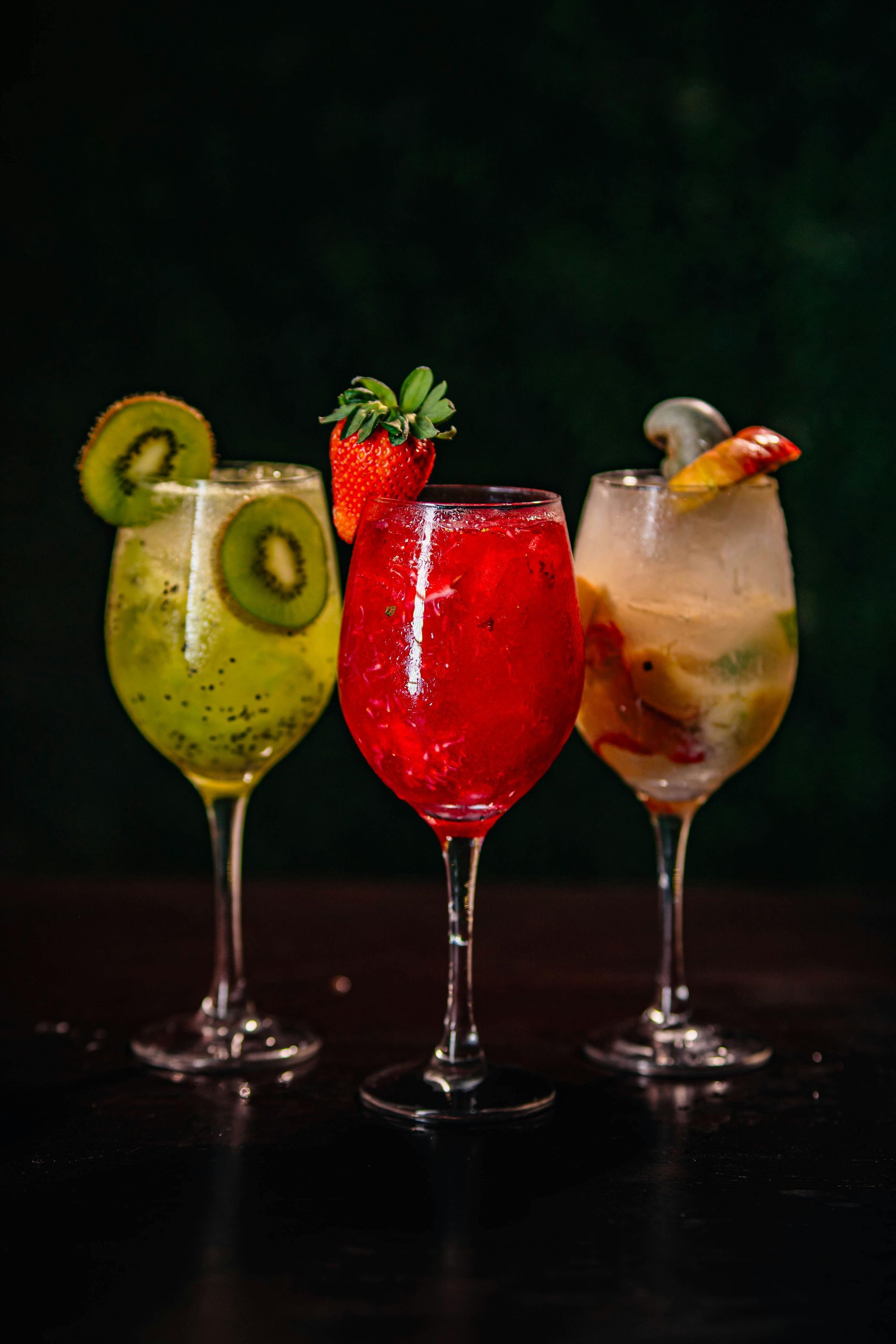 Three colorful fruit cocktails on a dark table, garnished with kiwi, strawberry, and citrus.