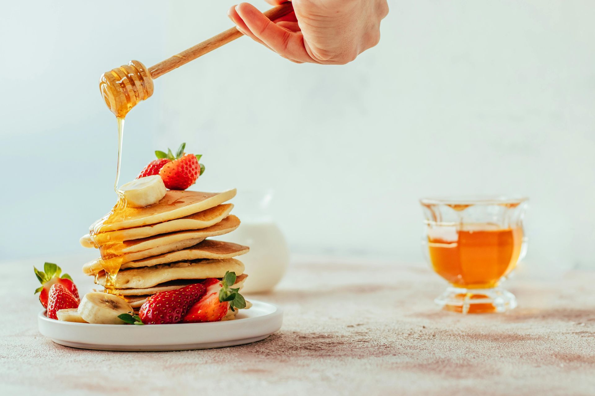Hand pouring honey over a stack of pancakes topped with strawberries, with a cup of tea nearby.