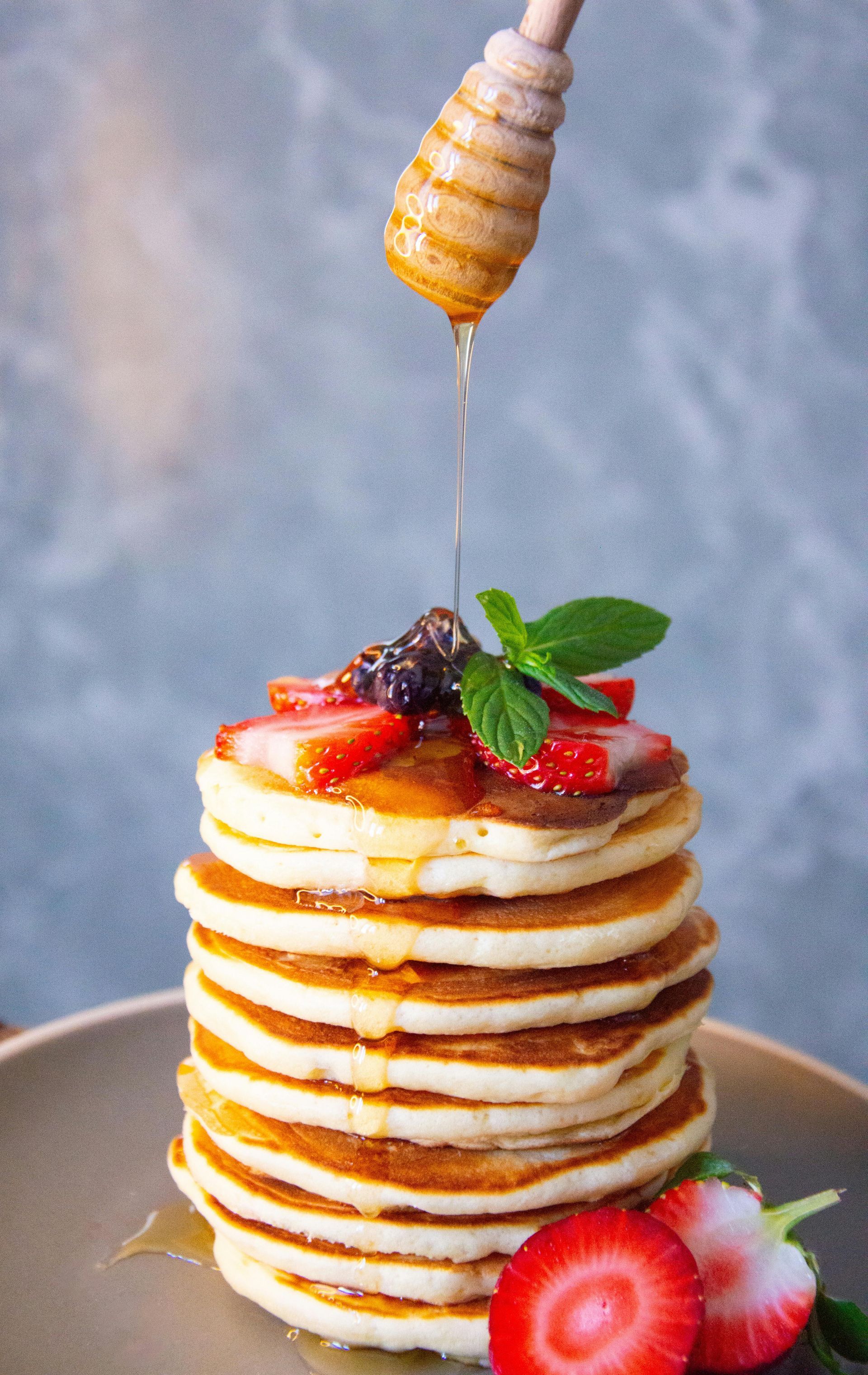 Stack of pancakes topped with strawberries and mint, with syrup being poured from above