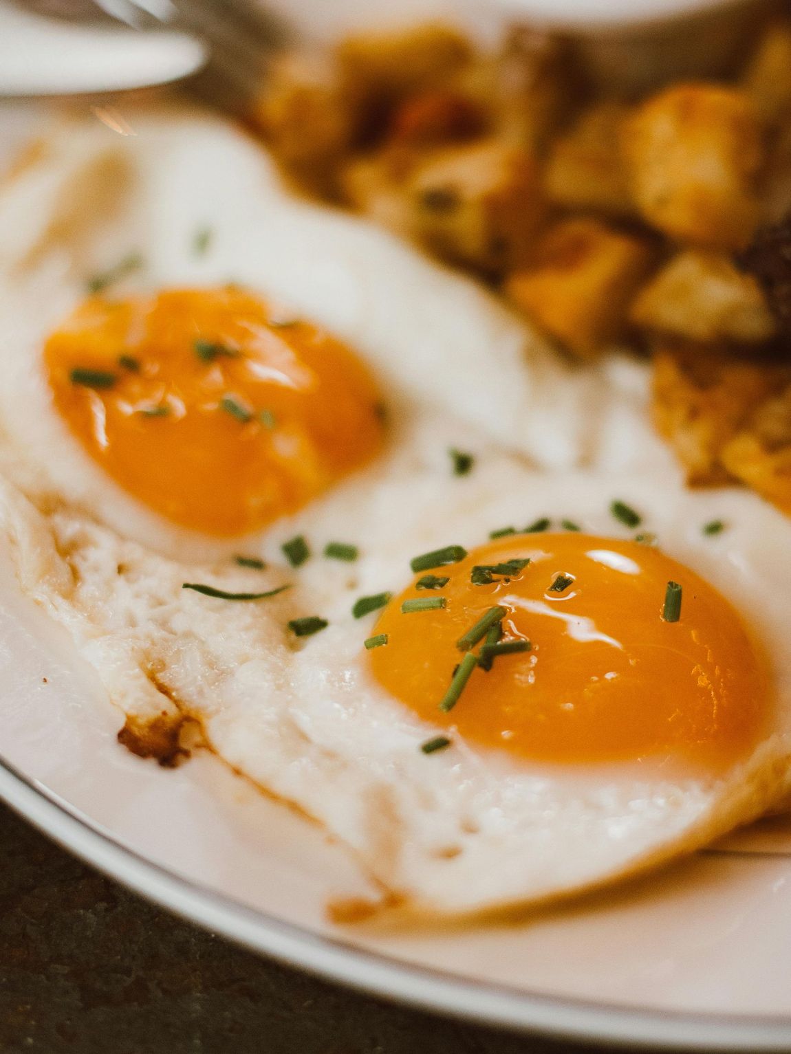 Two sunny-side-up eggs with herbs on a plate, with roasted potatoes in the background