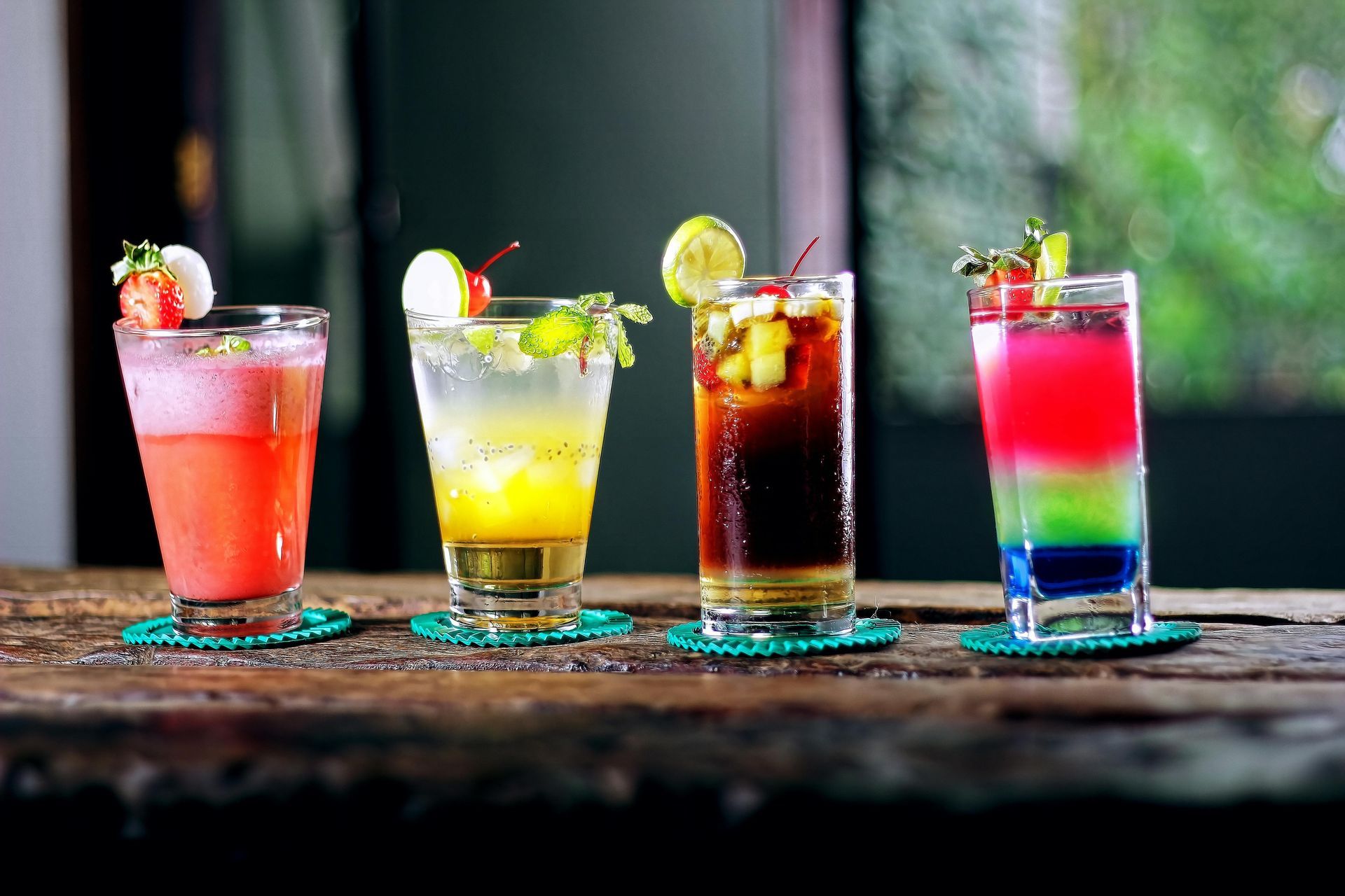Four colorful cocktails in glasses on a rustic table, garnished with fruit and ice.