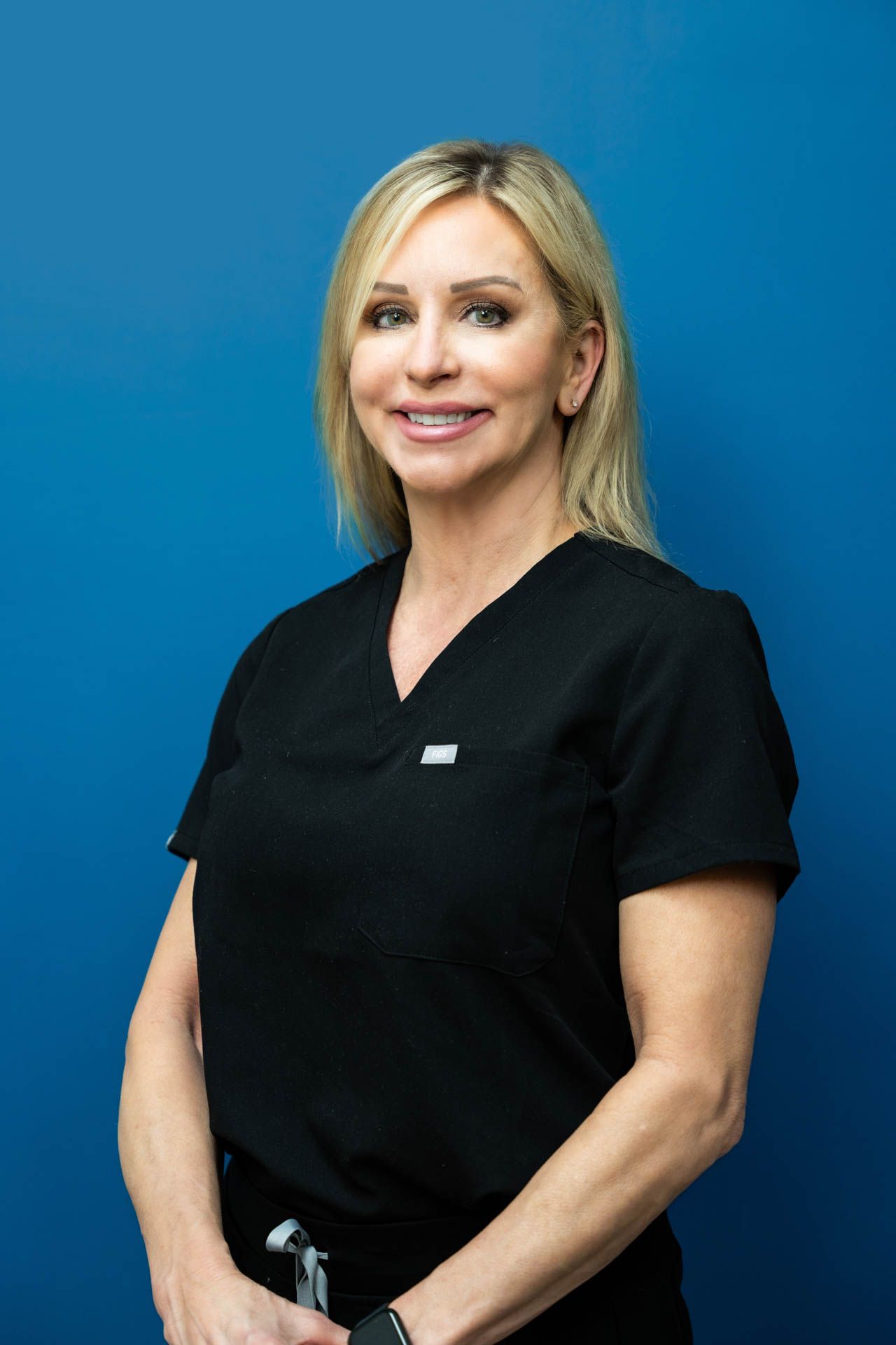A woman in a black scrub top is standing in front of a blue wall and smiling.