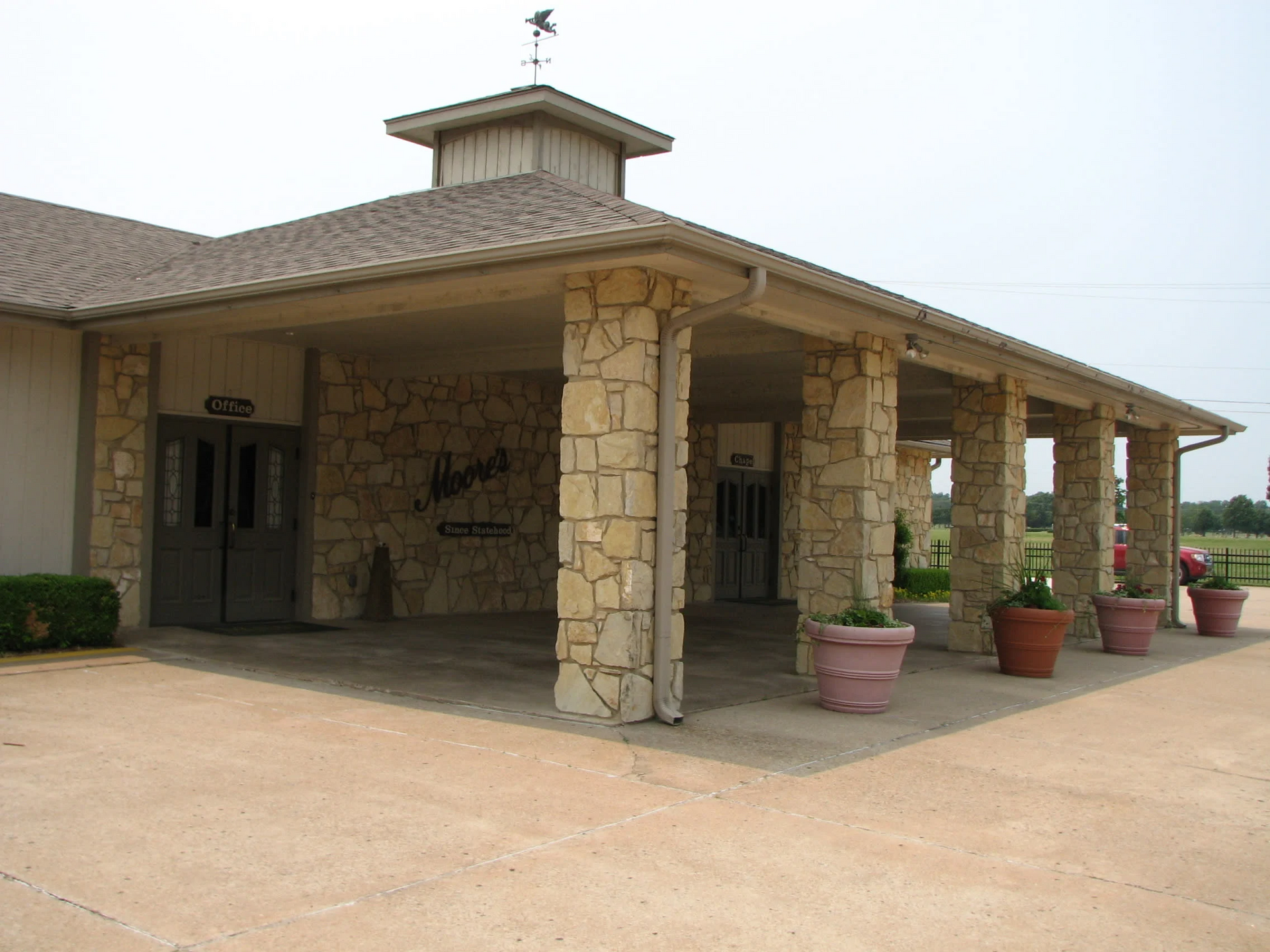 Stone building entrance with covered porch, tan roof, and potted plants along the driveway