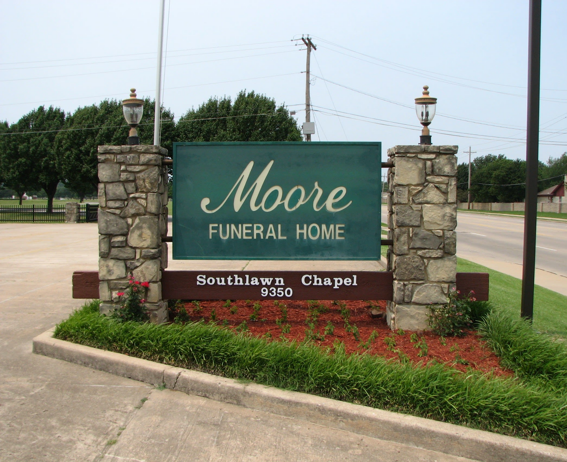 Moore Funeral Home sign on stone pillars beside a road and red flower bed