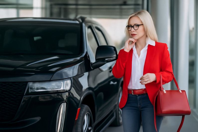 Woman in red blazer standing beside a black SUV, holding a phone and red handbag.