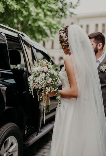 Bride in white veil holding a bouquet beside a black car, with groom standing behind her.