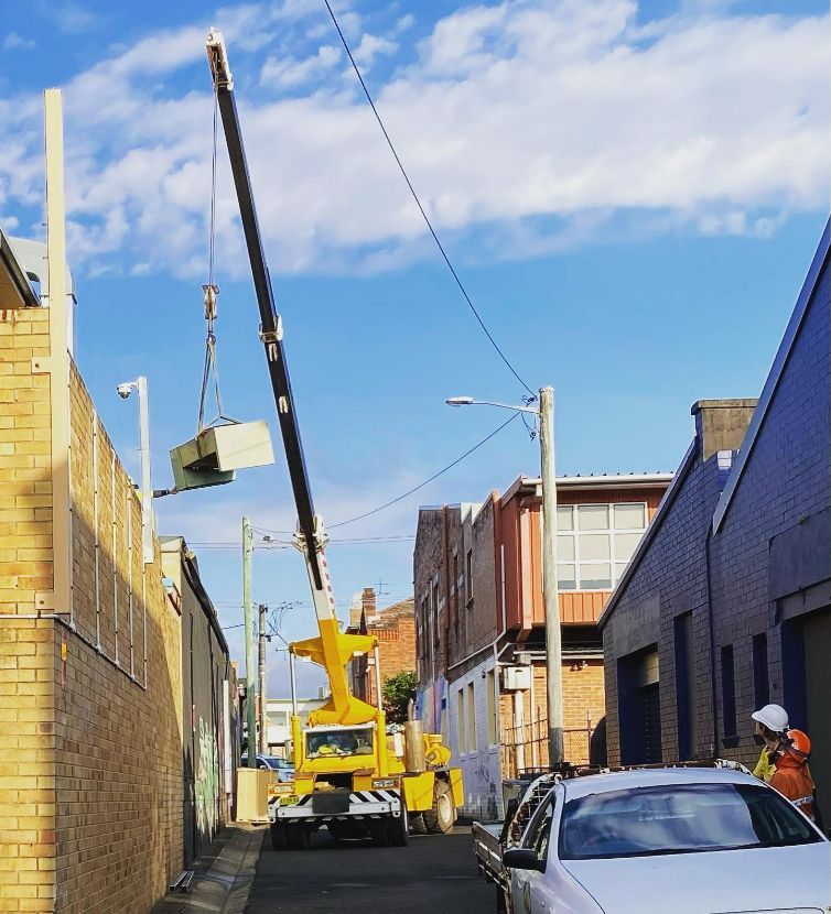 A Yellow Crane Is Lifting A Box In A Narrow Alleyway — Frigid-Air PTY Ltd In Ballina, NSW