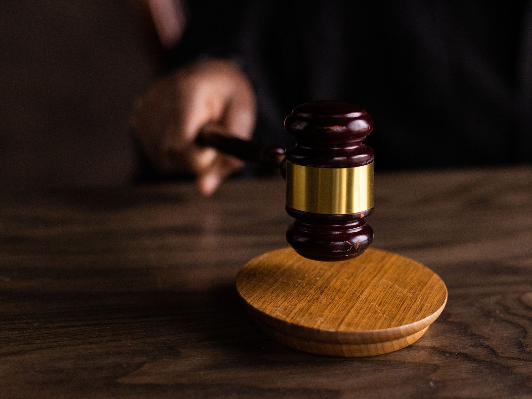 A judge's gavel with a gold band is captured mid-motion above a wooden sound block on a dark wooden table.