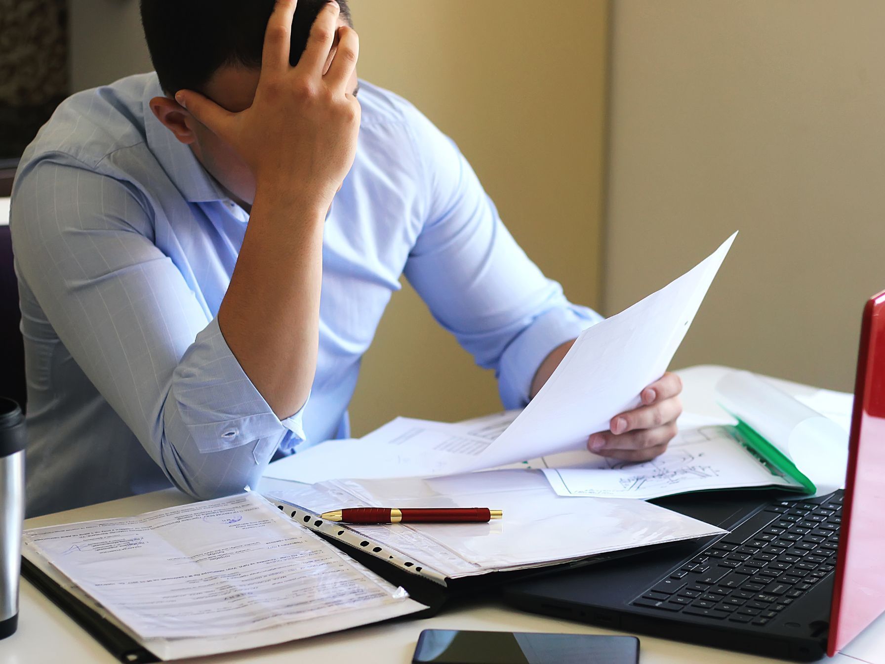 A stressed man in a light blue shirt holds a document while surrounded by paperwork, a laptop, and office supplies.