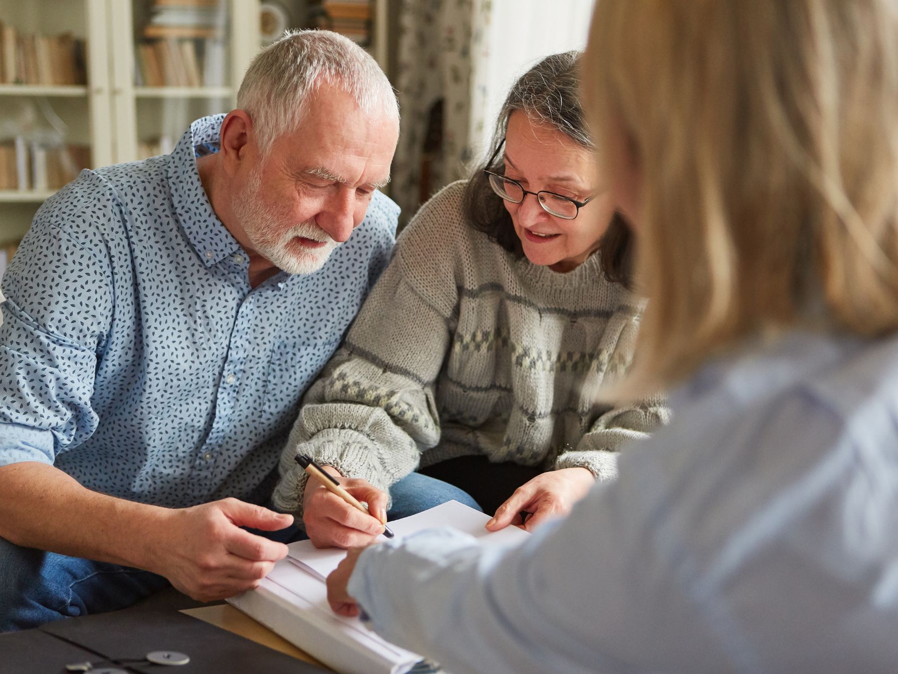 An elderly couple smiles while reviewing and signing documents with a professional in a cozy home setting.