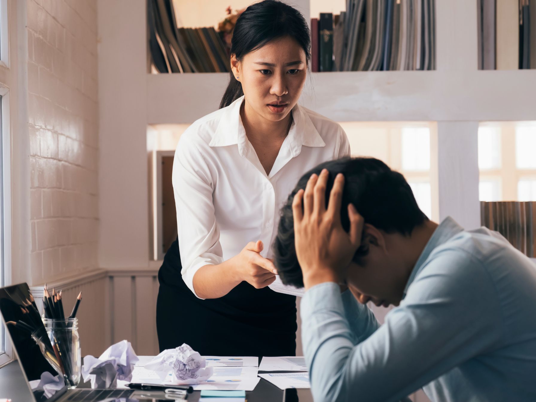 A frustrated woman in business attire confronts a stressed man at a cluttered desk filled with papers and charts.