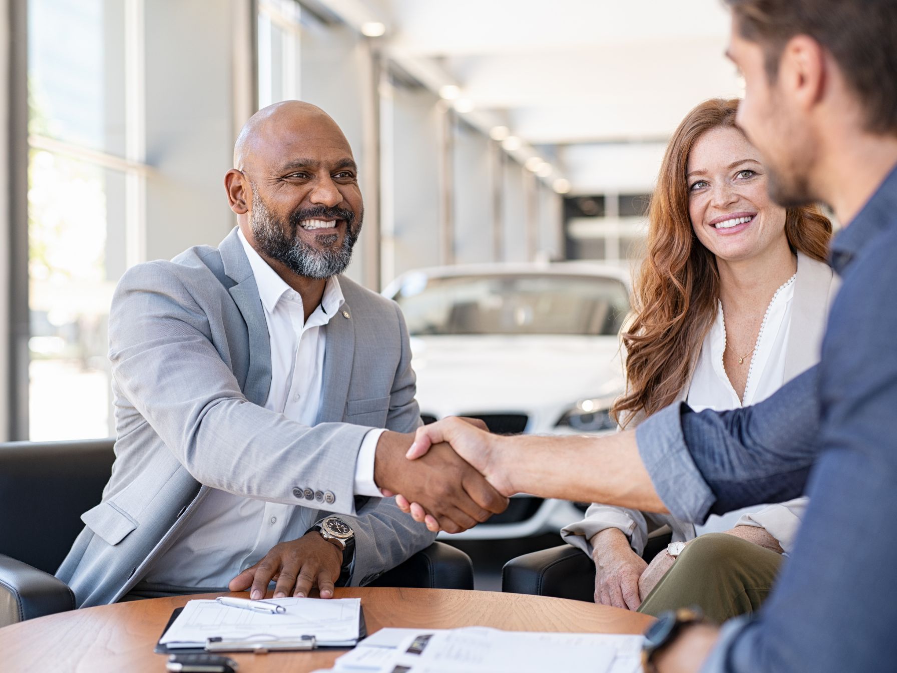 A smiling salesman in a light gray suit shakes hands with a male customer while a happy woman watches