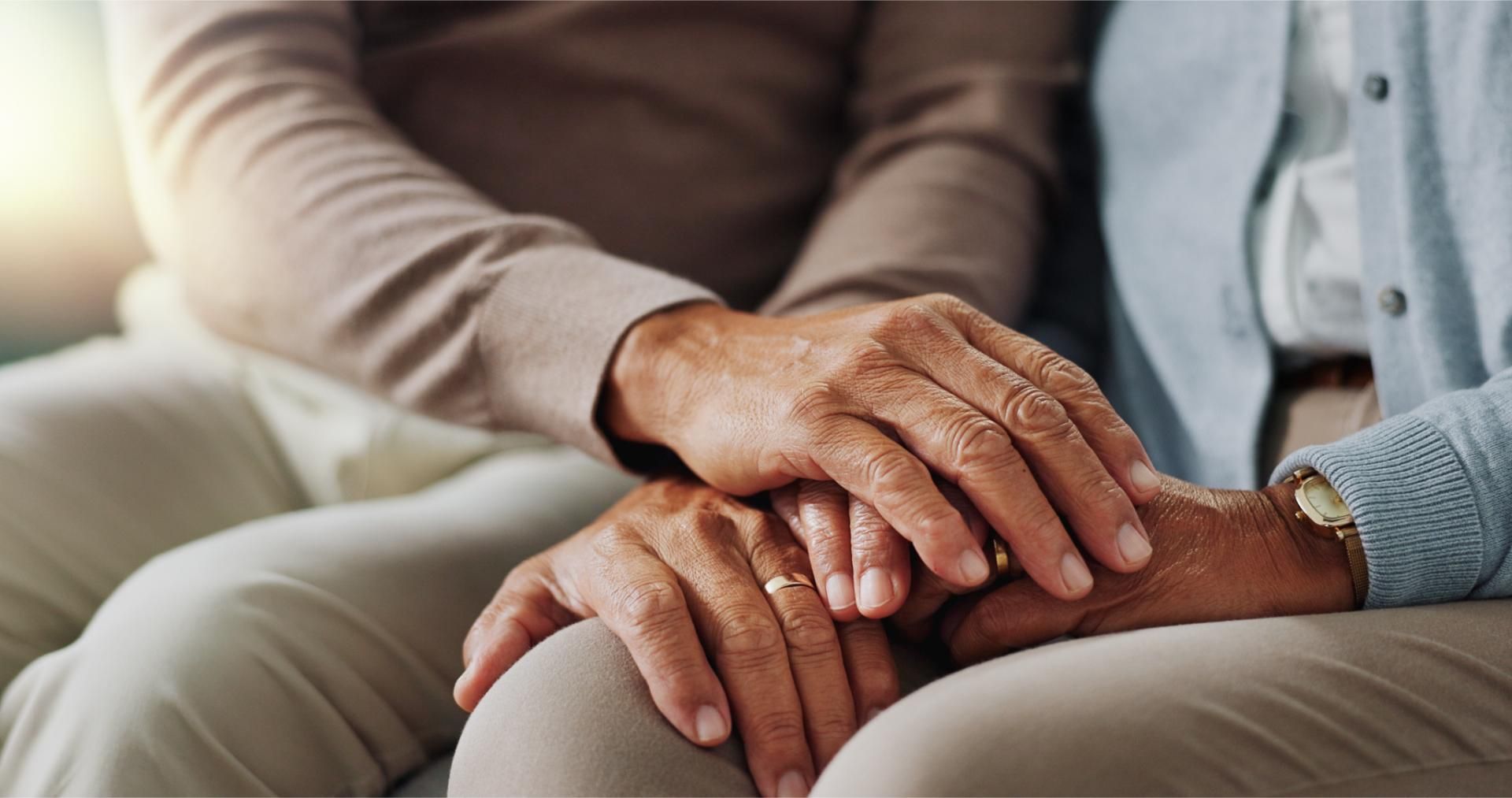An elderly couple is holding hands while sitting on a couch.