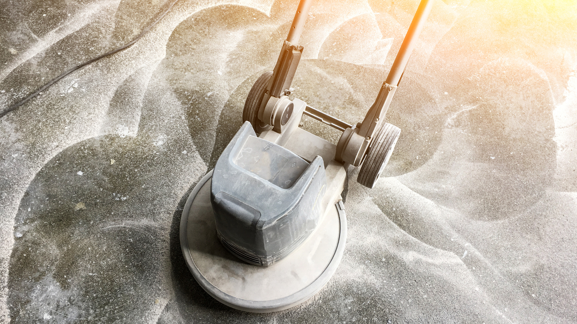 Floor buffer cleaning a concrete surface, dust visible, bright light.