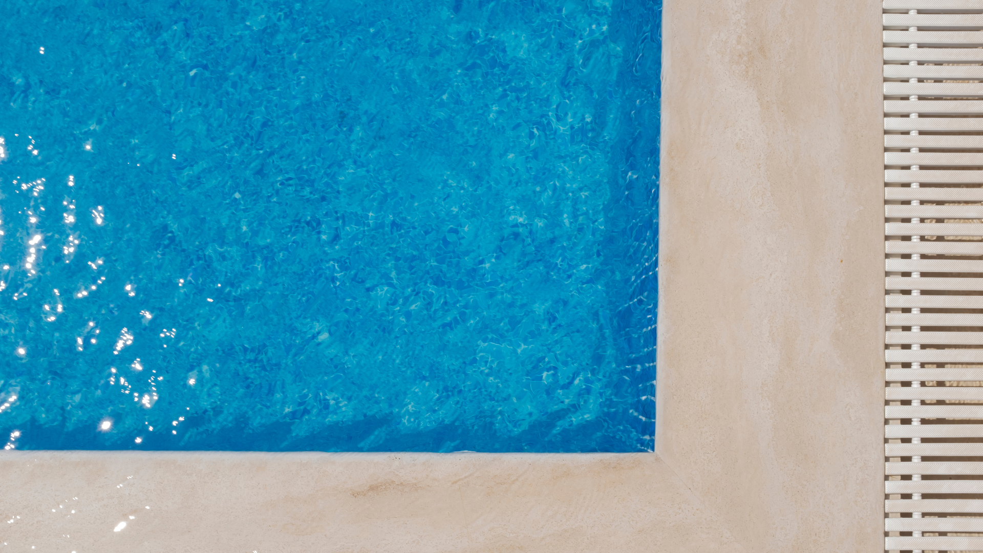 Blue swimming pool corner next to beige deck and white grate.