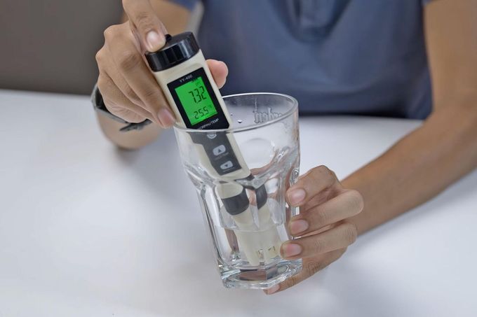 Person testing water with a digital pH meter in a clear glass on a white table