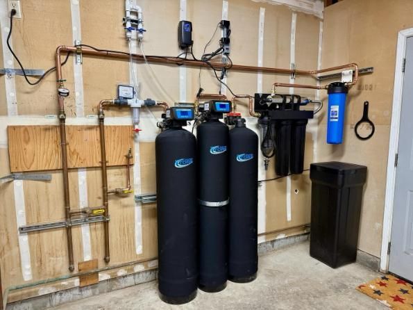 Water treatment equipment in a garage: three black tanks, blue filter unit, and exposed plumbing on the wall
