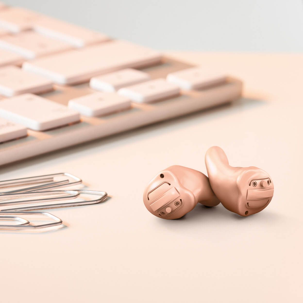 Two flesh-toned hearing aids next to a light pink keyboard and paperclips on a table.
