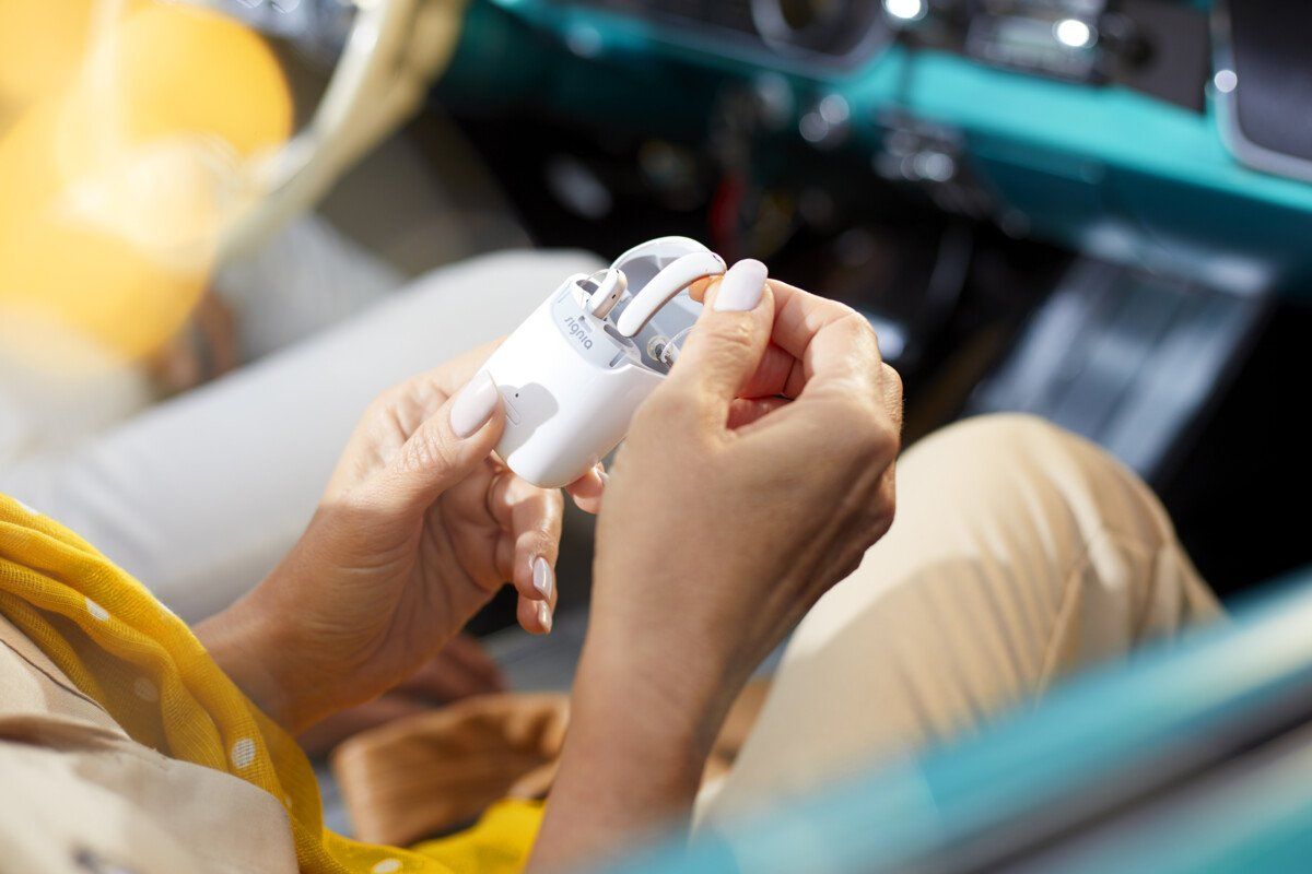 Hands holding a small white object, possibly a hearing aid, inside a vintage car.