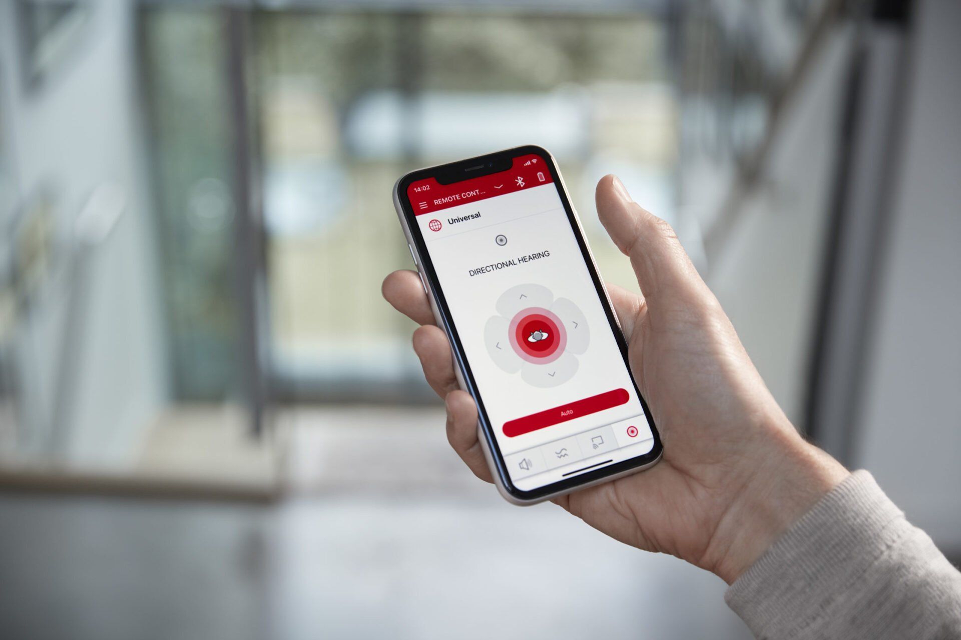 Person holding a smartphone displaying hearing aid app with a circular graphic and text; a blurred indoor setting.