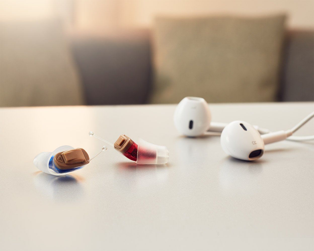 A broken hearing aid and white earbuds on a table, soft-focus background of couch.