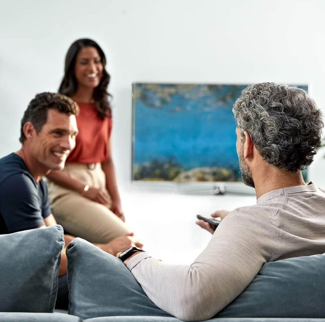 Three people watching TV, smiling. Two sit on the sofa, one holding a remote. TV on the wall.
