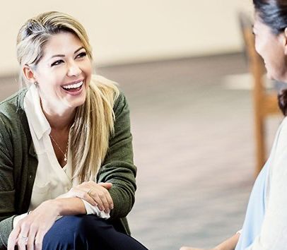 Woman laughing during conversation. Sitting indoors, light-colored walls. She wears a cardigan, white shirt, and dark pants.