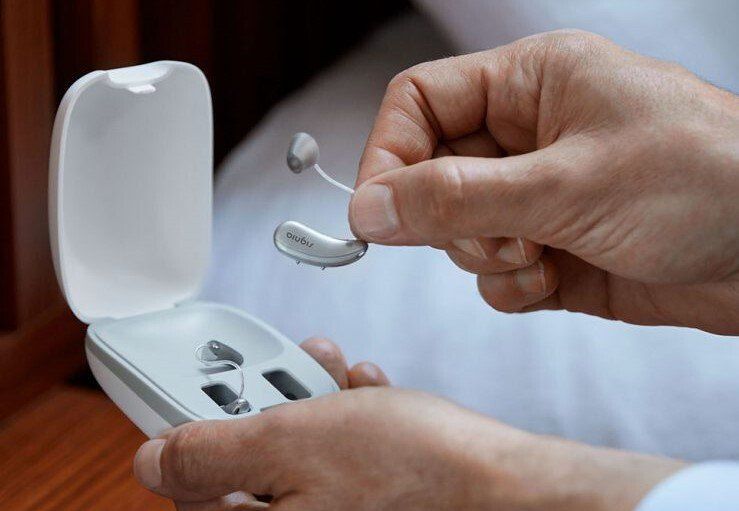 Hands holding hearing aids near an open charging case, bedroom setting.
