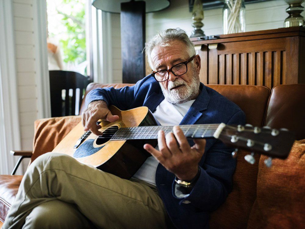 Older person with glasses playing acoustic guitar on a couch in a living room.