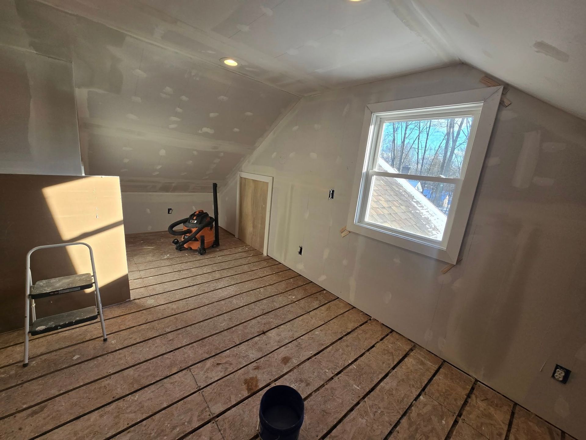 Interior of an attic under construction. Drywall, wood flooring, window, ladder, and tools are visible.