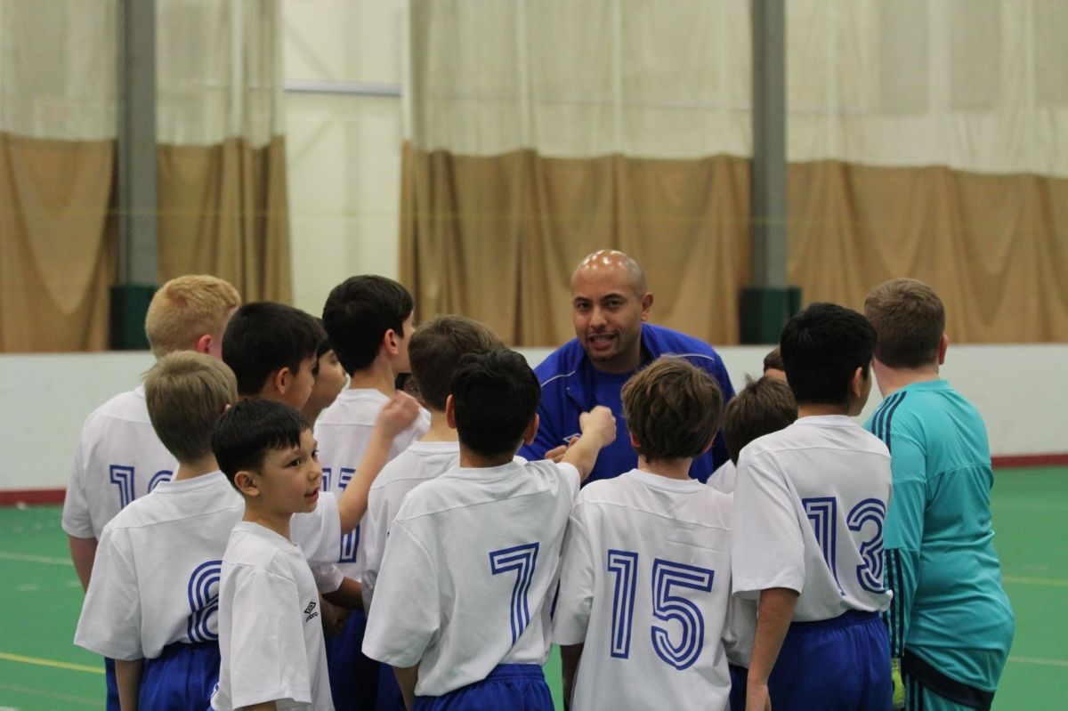 A coach in a blue shirt gathers a team of youth soccer players in white jerseys for a huddle on an indoor field.