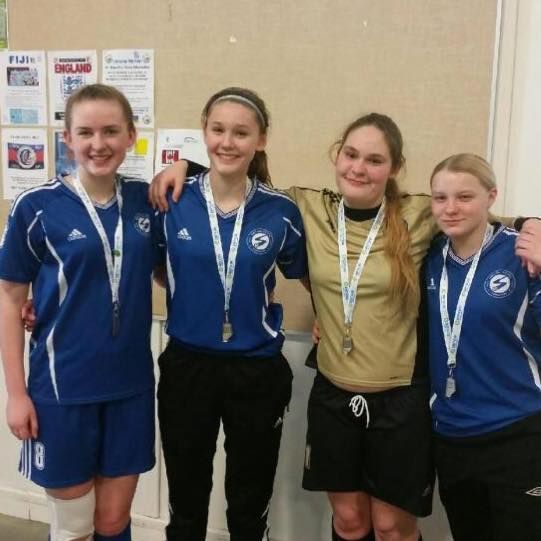 Four people in sports uniforms with medals around their necks stand side-by-side, smiling indoors against a bulletin board.