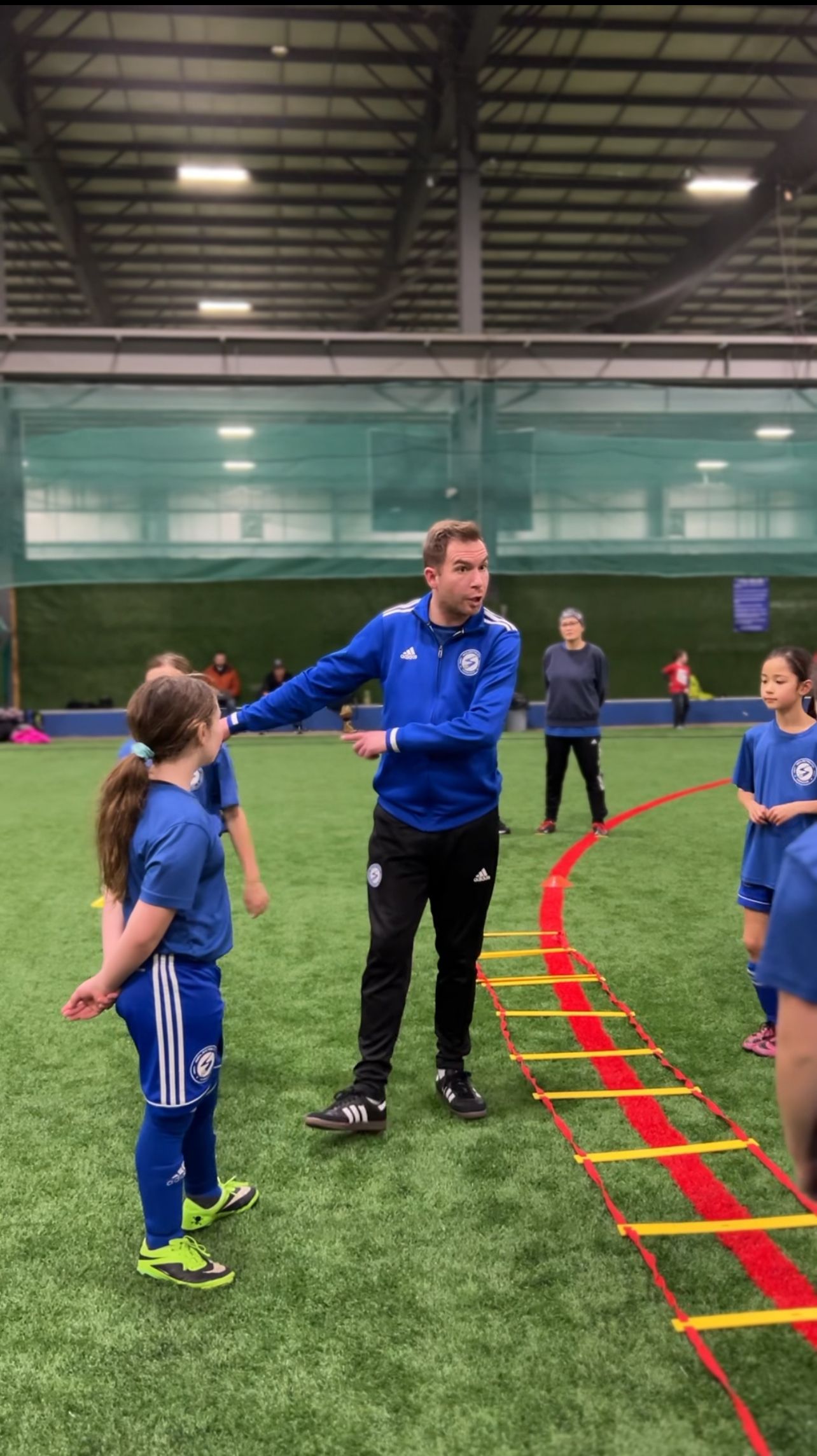 A coach in a blue jacket instructs players on an indoor soccer field with an agility ladder on the turf.