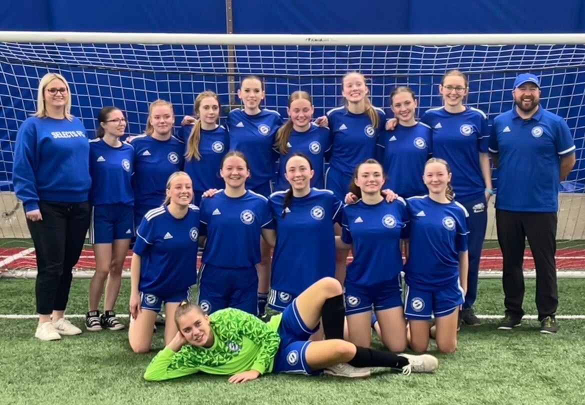 A team of soccer players in matching blue uniforms and two coaches pose for a photo on an indoor field with blue netting.