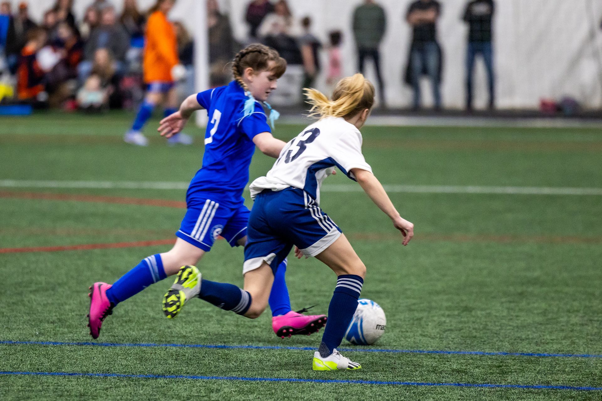 Two soccer players compete for the ball on an artificial turf field, one in a blue uniform and one in a white jersey.