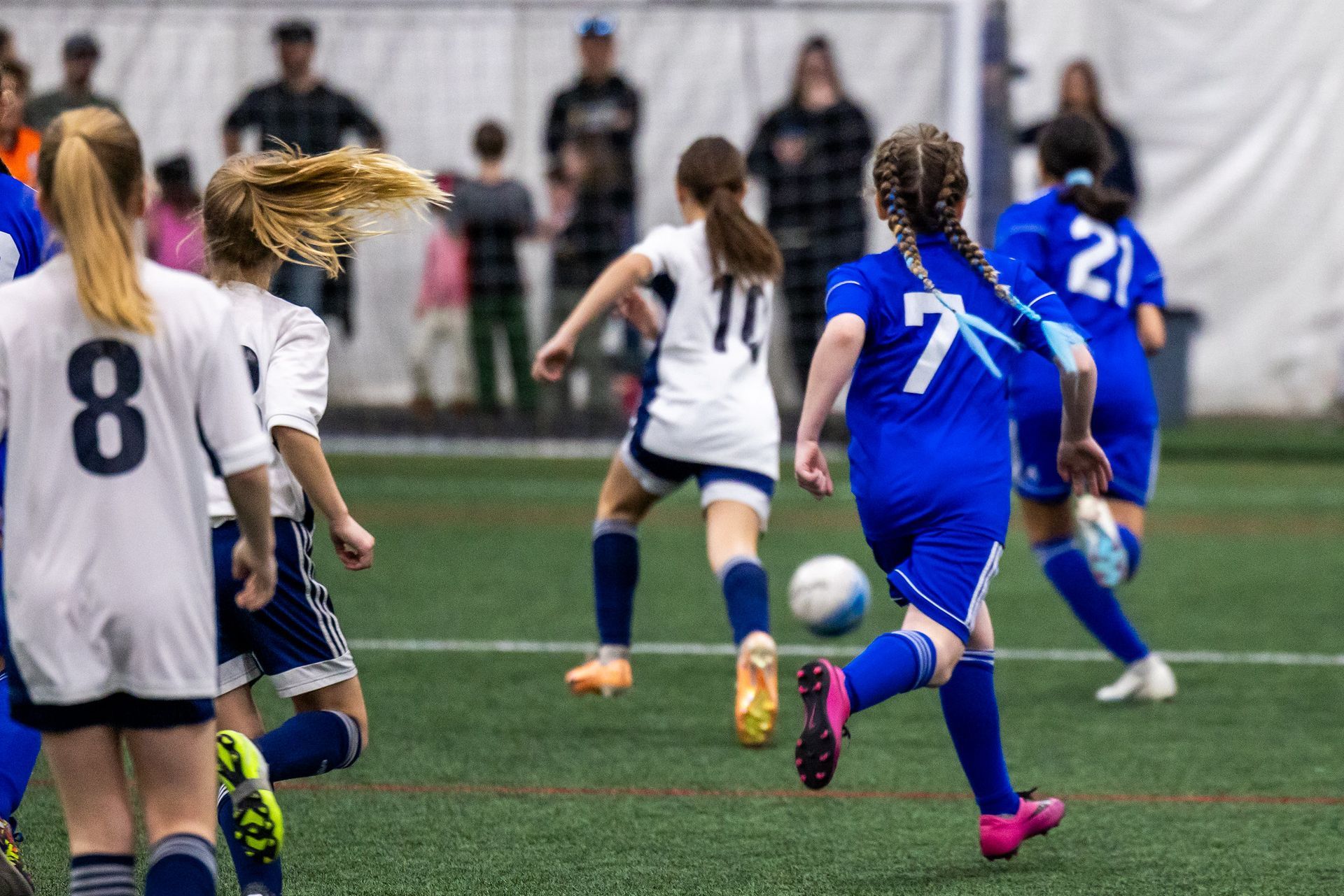 Youth soccer players in white and blue uniforms running on an indoor turf field during a game.
