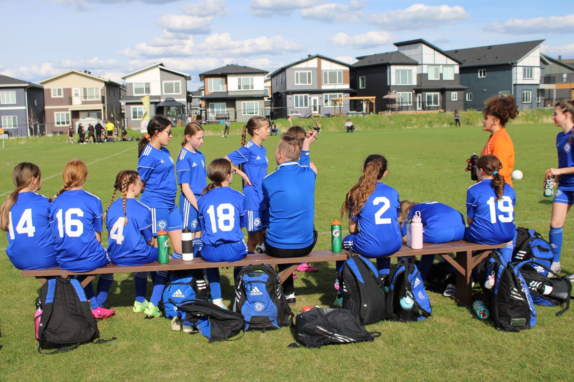 Soccer team in blue jerseys sitting on a bench on a grassy field in front of suburban houses.