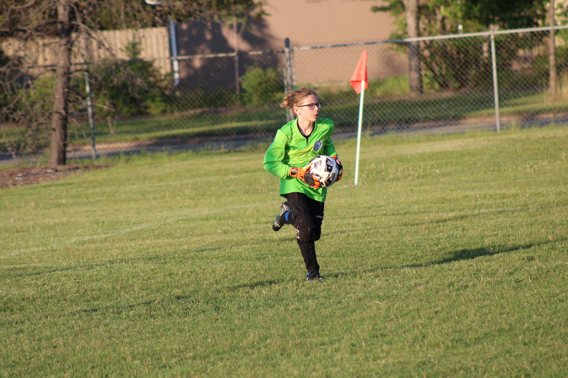 A soccer goalie in a neon green jersey runs across a grass field holding a soccer ball near a corner flag.