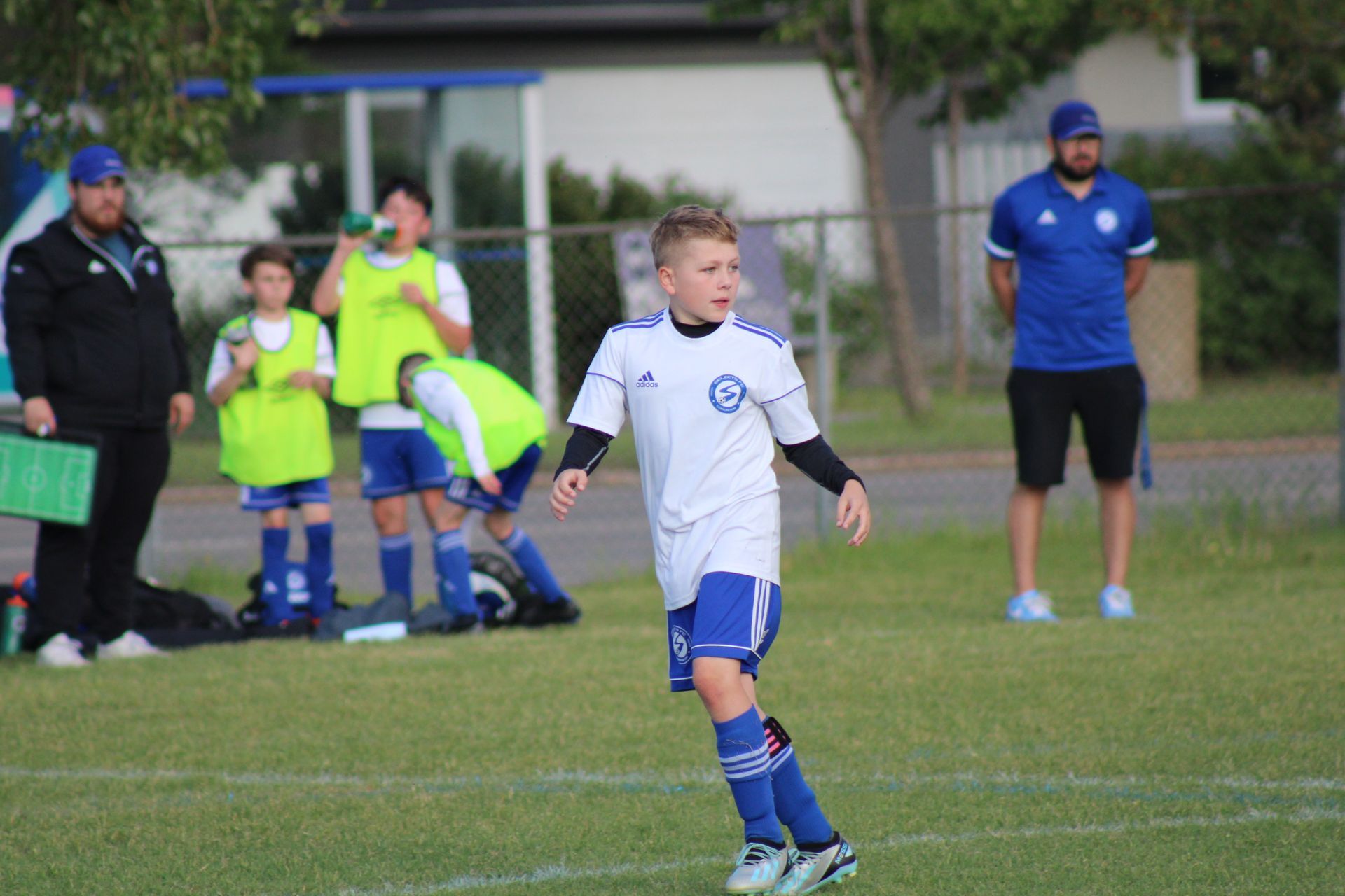 A soccer player in a white jersey and blue shorts stands on a grass field during a practice with coaches and teammates.
