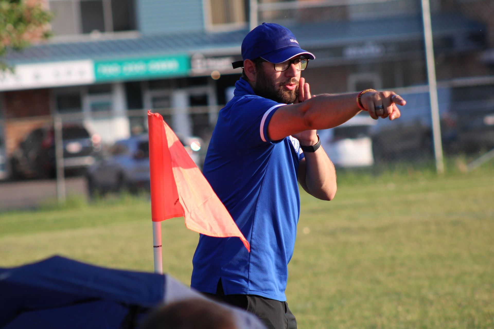 A coach in a blue shirt and cap stands on a field, pointing and gesturing toward the distance next to a red flag.