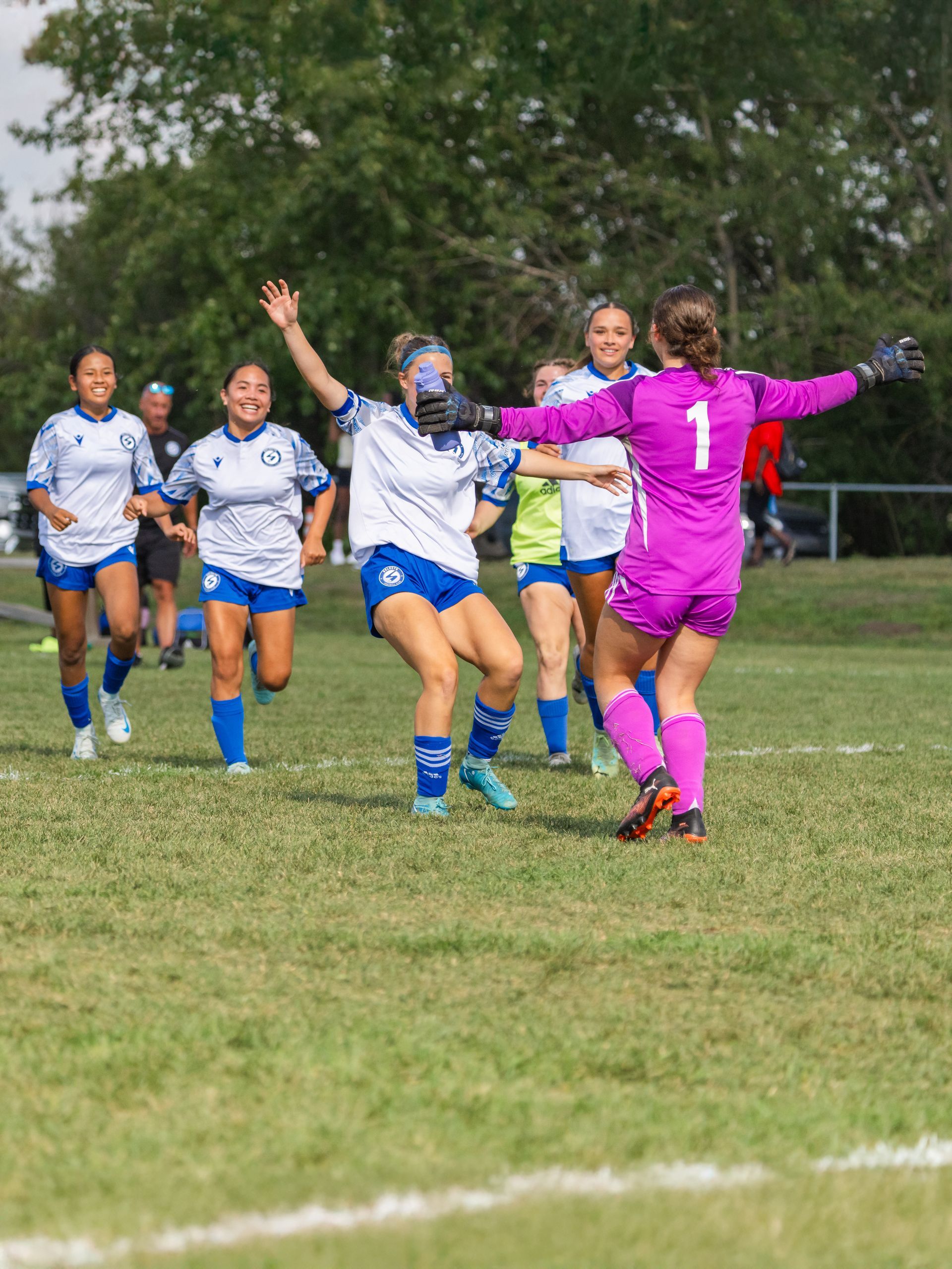 Soccer players in white and blue uniforms celebrate on a field with their teammate, a goalkeeper in a magenta jersey.