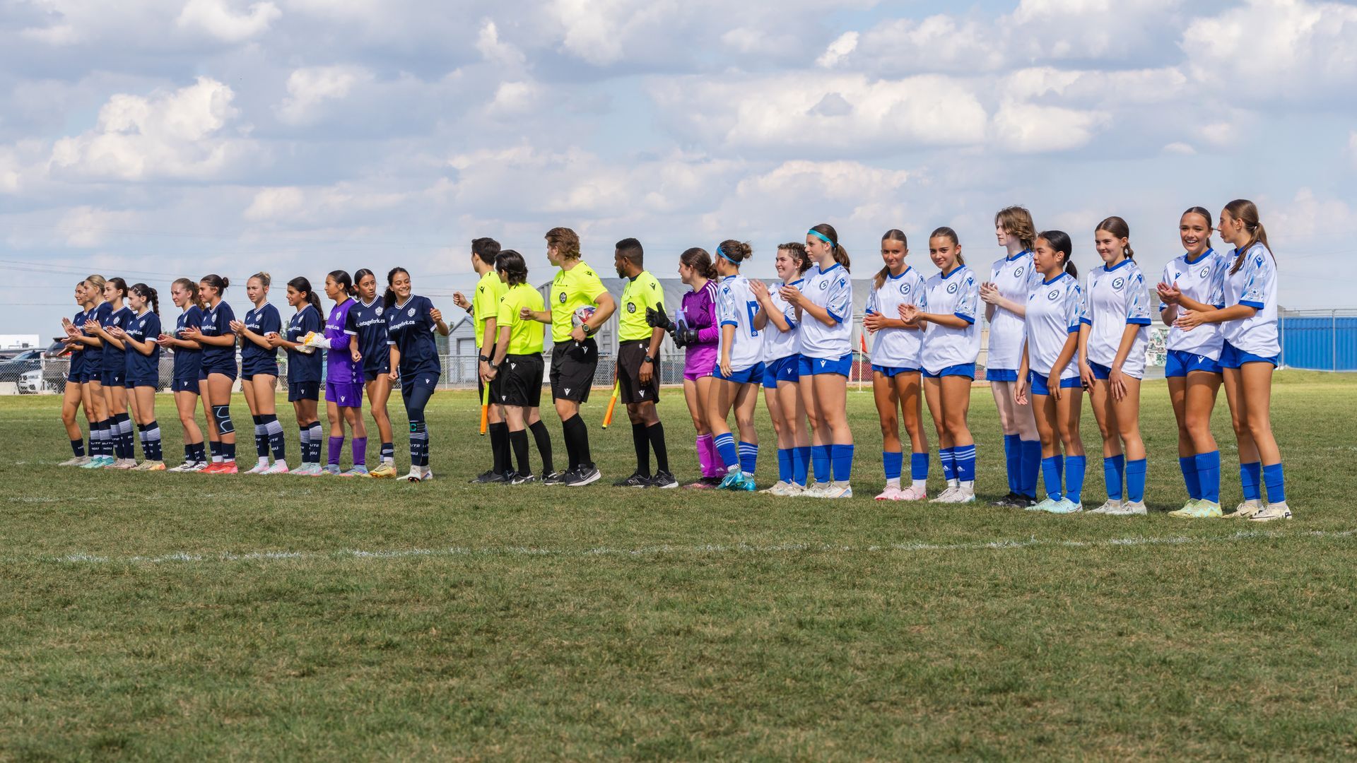 Two teams of soccer players in blue and white jerseys line up on a grassy field with match officials.