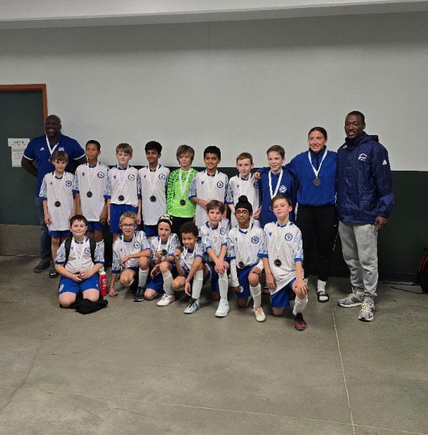 A youth soccer team in white and blue uniforms poses with medals alongside two coaches in an indoor hallway.